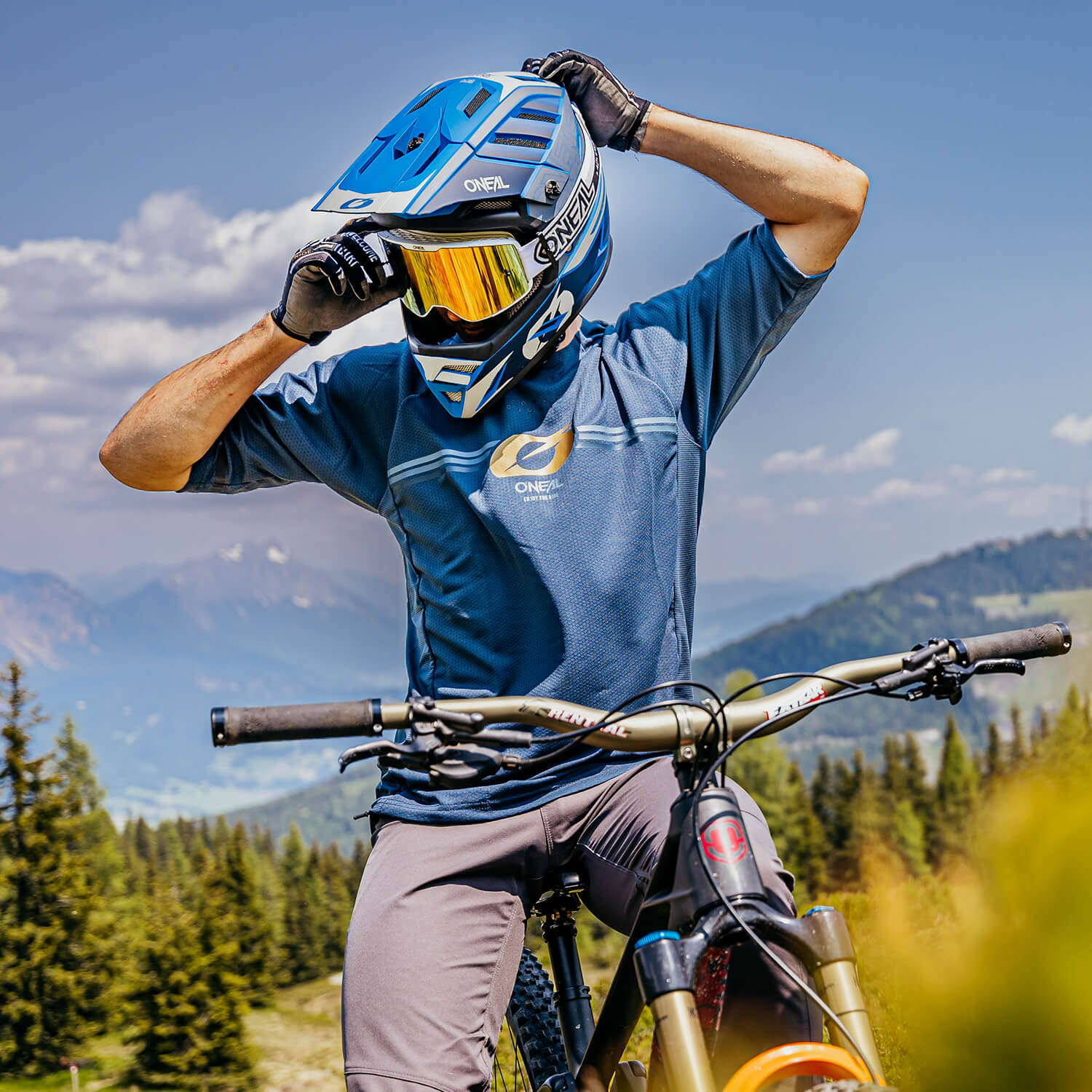 Ein Mountainbiker mit blauem Helm, reflektierender Schutzbrille und blauem Trikot passt seine Ausrüstung an, während er mit seinem Fahrrad auf einem malerischen Pfad steht. Im Hintergrund sind Berge, Bäume und ein klarer Himmel mit vereinzelten Wolken zu sehen.