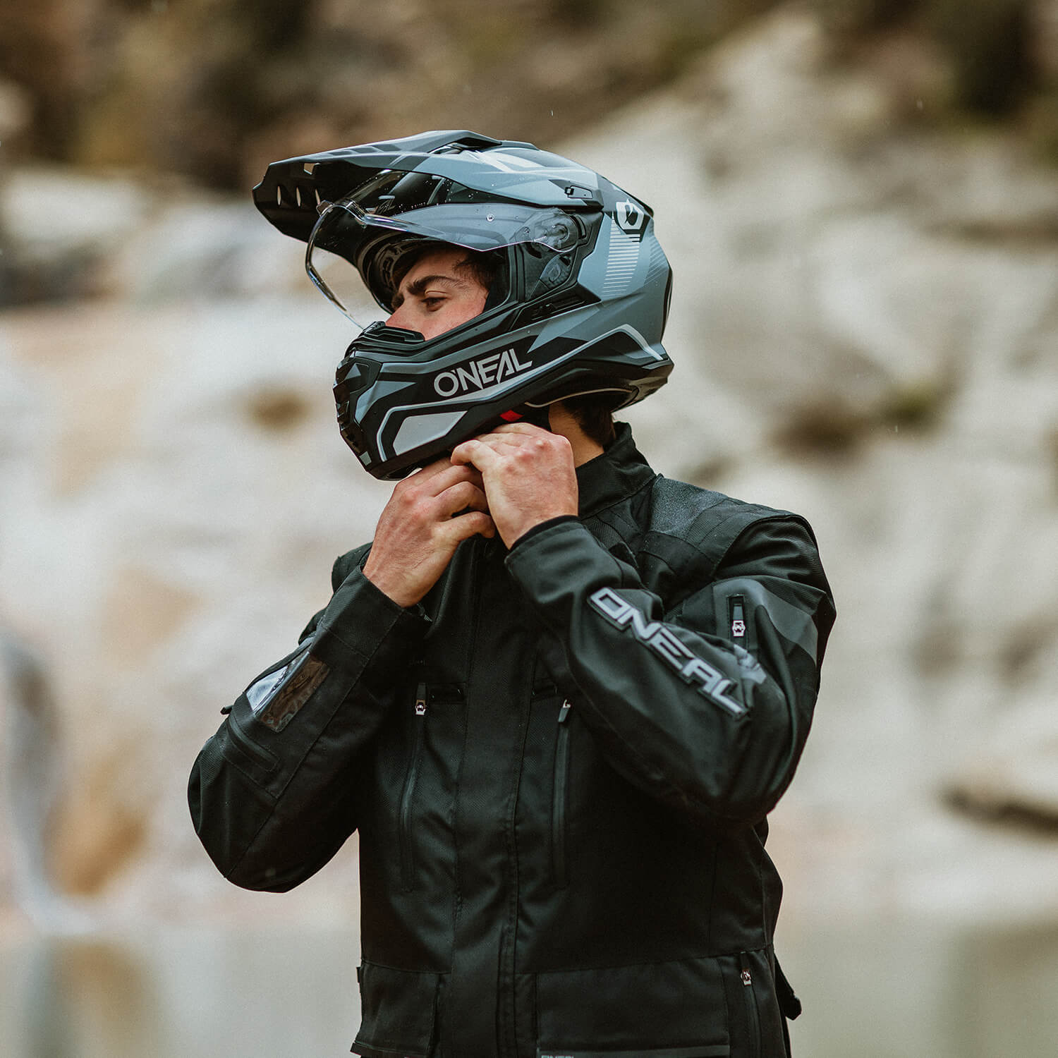 Eine Person mit einem schwarz-grauen O'Neal-Motorradhelm und einer schwarzen Jacke passt den Helmgurt an. Der Hintergrund ist eine verschwommene Außenkulisse mit natürlichem Licht.