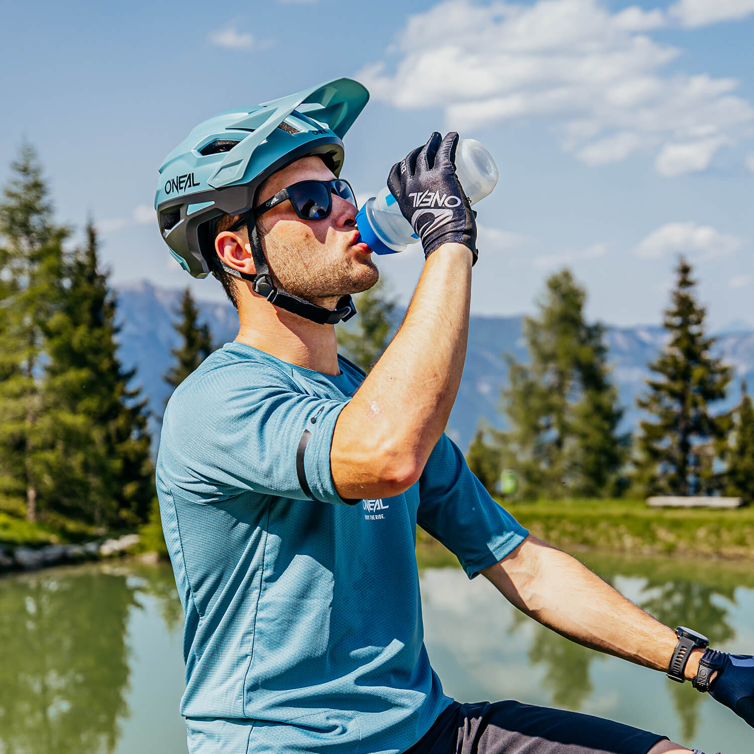 Ein Radfahrer mit blauem Helm, Sonnenbrille, blauem Trikot und Handschuhen trinkt aus einer Wasserflasche. Er sitzt vor einer malerischen Kulisse mit Bäumen, einem ruhigen See und Bergen unter einem blauen Himmel.