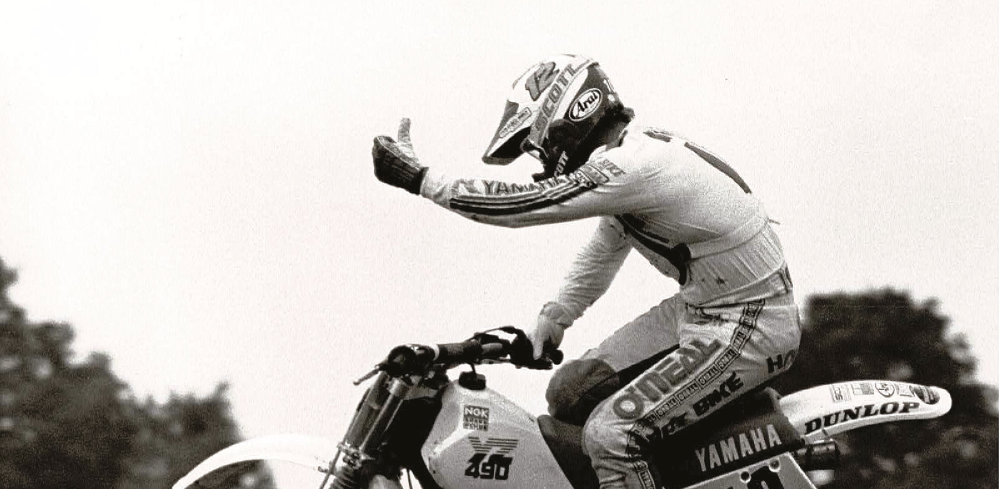 Eine Person auf einem Yamaha-Motocross-Motorrad, in der Luft während eines Sprungs. Der Fahrer trägt die komplette Rennmontur und einen Helm, mit ausgestrecktem linken Arm und erhobenem Daumen. Im Hintergrund sind verschwommene Bäume zu sehen, die auf Bewegung hindeuten.