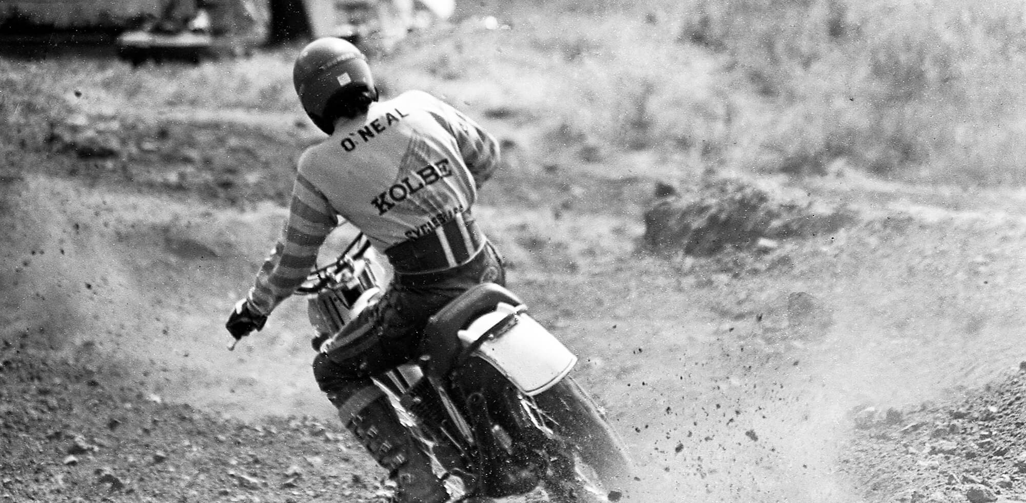 Schwarzweißbild eines Motorradrennfahrers, der einen Feldweg befährt. Der Fahrer, der einen Helm und ein Trikot mit der Aufschrift „O'NEAL“ auf dem Rücken trägt, wirbelt Staub auf, als er sich in eine Kurve legt. Der Hintergrund ist verschwommen, wodurch die Bewegung und Geschwindigkeit der Szene hervorgehoben werden.