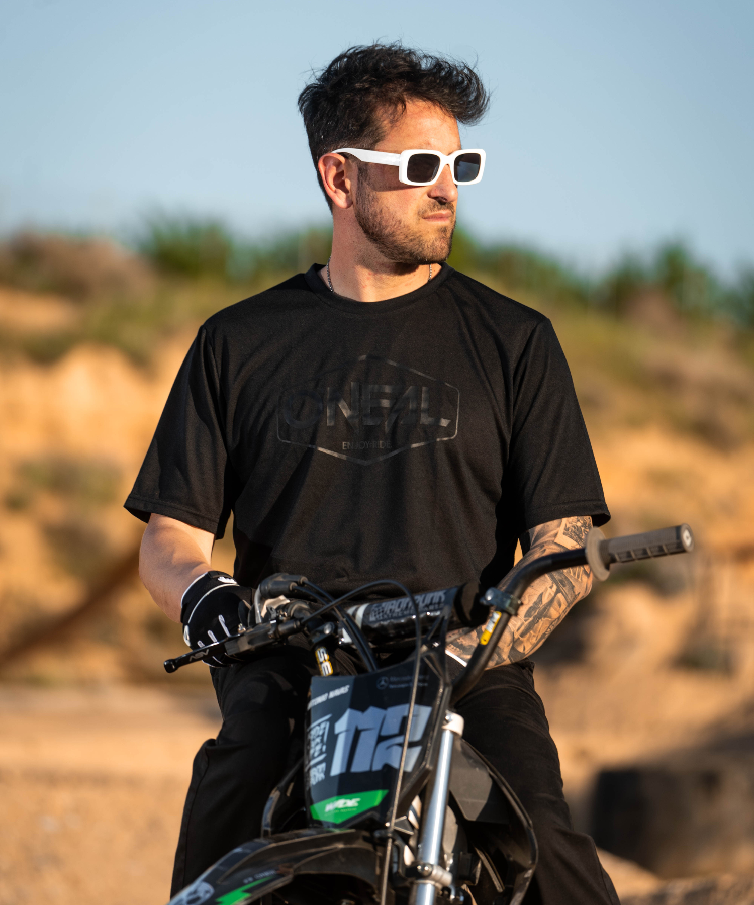 Ein Mann mit weißer Sonnenbrille und schwarzem T-Shirt sitzt auf einem Motocross-Motorrad im Freien und schaut zur Seite. Der Hintergrund ist unscharf mit sandigem Gelände und Grünzeug.