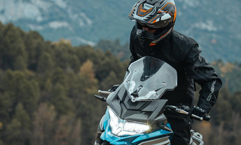 Eine Person mit Helm und schwarzer Jacke fährt mit einem Motorrad durch eine bergige Gegend mit dichten grünen Bäumen im Hintergrund.
