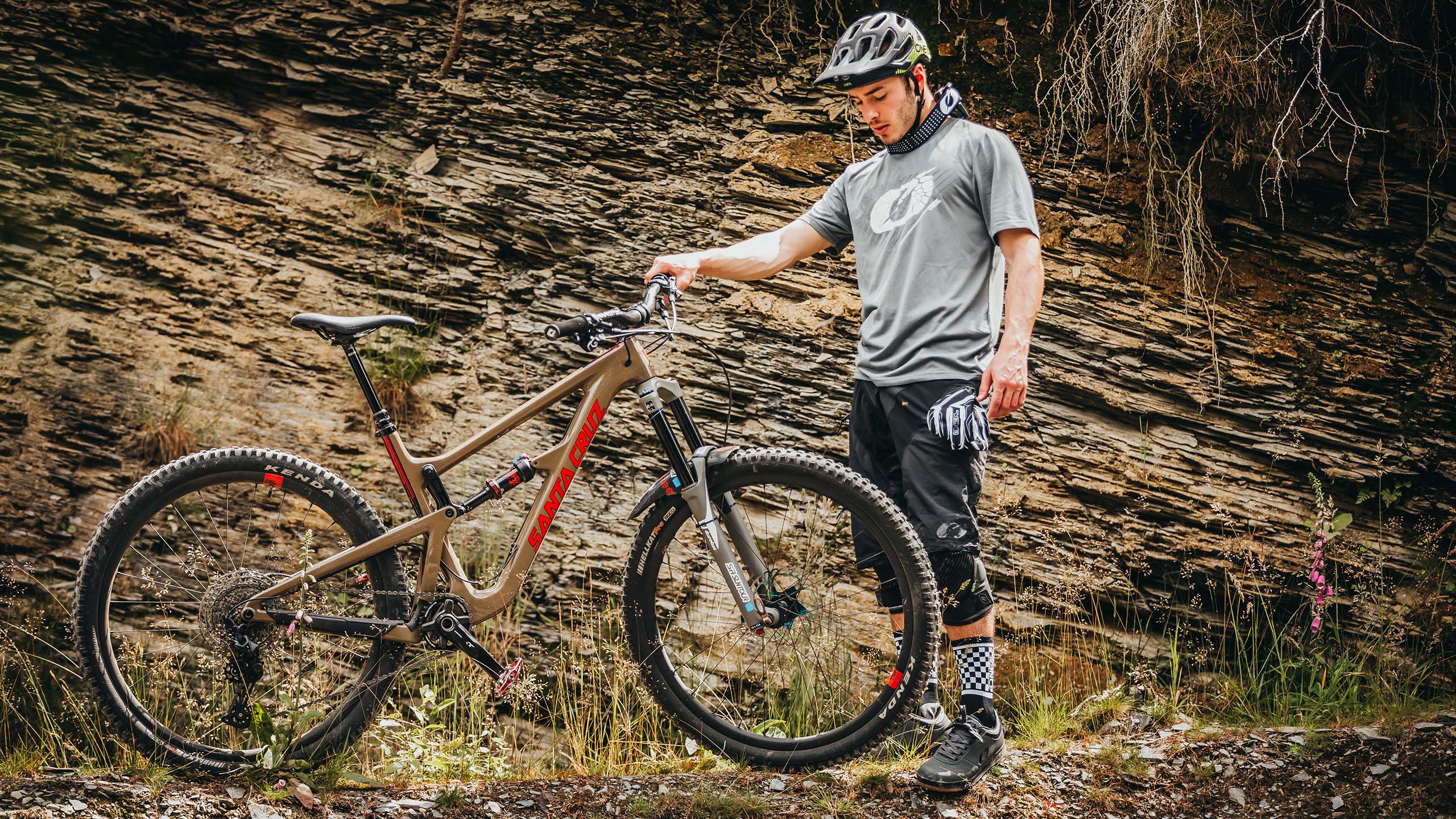Eine Person in Fahrradkleidung steht neben einem Mountainbike auf einem felsigen Pfad. Sie trägt einen Helm, ein graues Hemd, schwarze Shorts, Knieschützer und karierte Socken. Im Hintergrund sind eine schroffe Felswand und Grünflächen zu sehen.