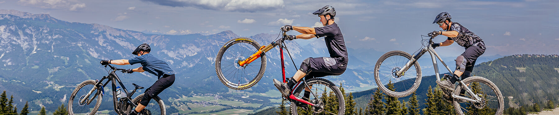 Drei Radfahrer führen vor der Kulisse eines bergigen Geländes und eines teilweise bewölkten Himmels Stunts auf Mountainbikes vor. Sie befinden sich in der Luft und demonstrieren Beweglichkeit und Gleichgewicht inmitten der weitläufigen Naturlandschaft.
