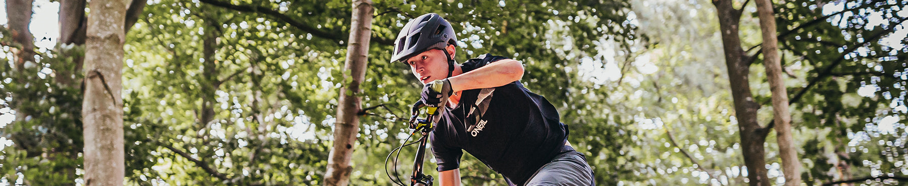 Ein Mountainbiker mit Helm und Handschuhen fährt auf einem Waldweg, umgeben von hohen Bäumen und durch deren Blätter gesprenkeltes Sonnenlicht fällt.