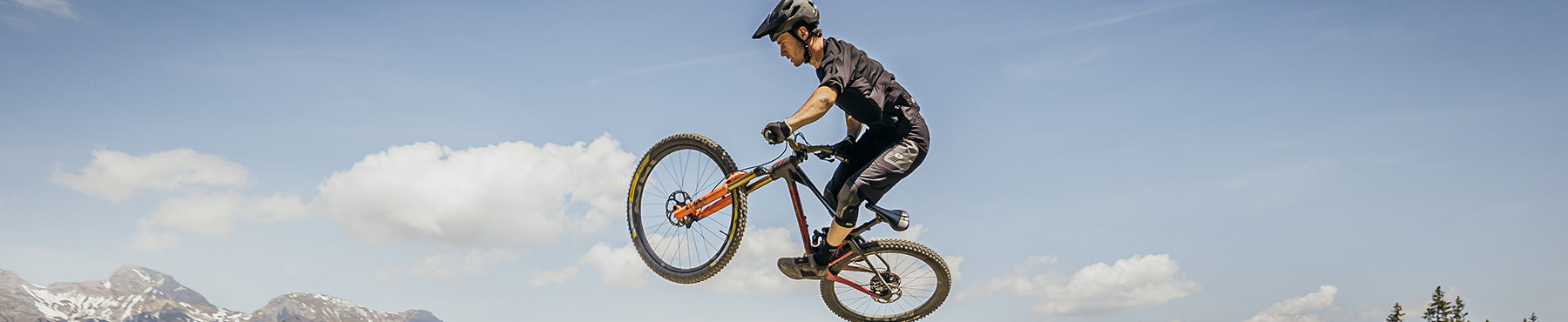 Eine Person mit Helm macht vor einem klaren blauen Himmel einen Sprung auf einem Mountainbike. Im Hintergrund sind schneebedeckte Berge und flauschige Wolken zu sehen.
