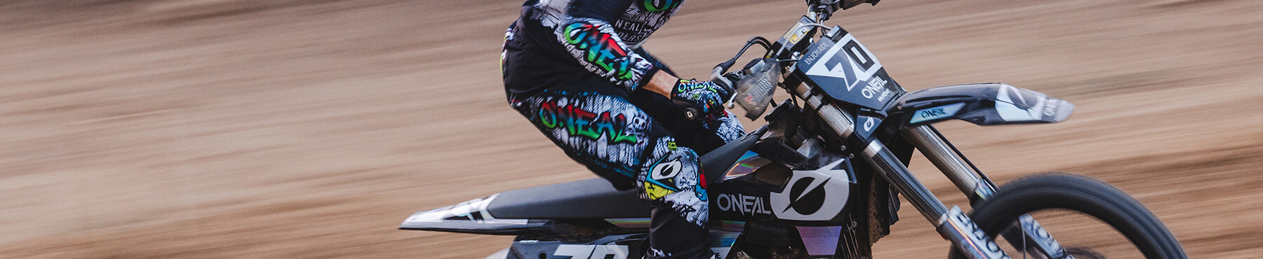 Eine Person fährt mit einem Dirtbike mit hoher Geschwindigkeit auf einer unbefestigten Strecke. Sie trägt farbenfrohe Rennausrüstung mit grafischen Mustern, darunter einen Helm und Handschuhe. Der Hintergrund ist unscharf, was die Bewegung betont.