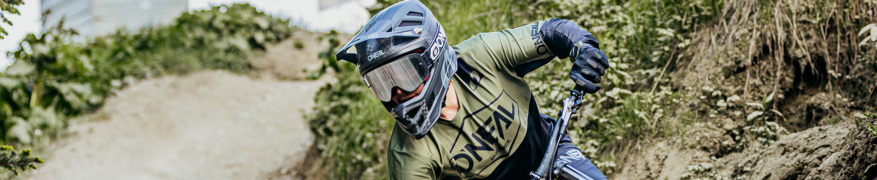Ein Biker mit Helm und Schutzbrille fährt auf einem von Grün umgebenen Feldweg bergab und konzentriert sich intensiv auf den vor ihm liegenden Weg.