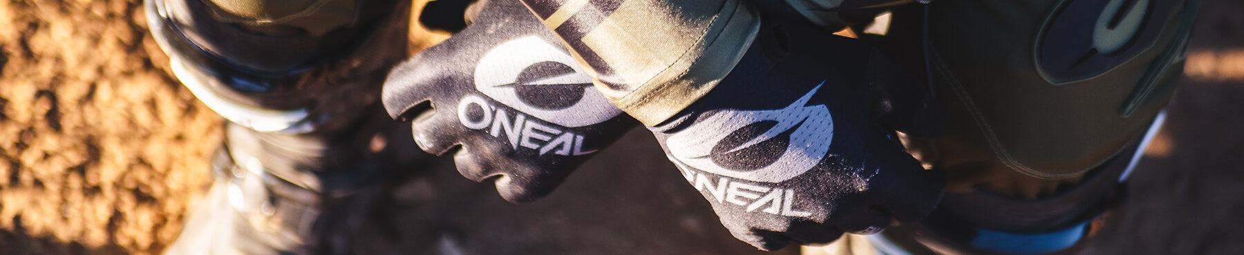 Nahaufnahme einer Person mit schwarzen O'Neal-Handschuhen und Motocross-Ausrüstung, die auf unbefestigtem Gelände hockt. Auf den Handschuhen ist das Logo der Marke deutlich zu sehen.