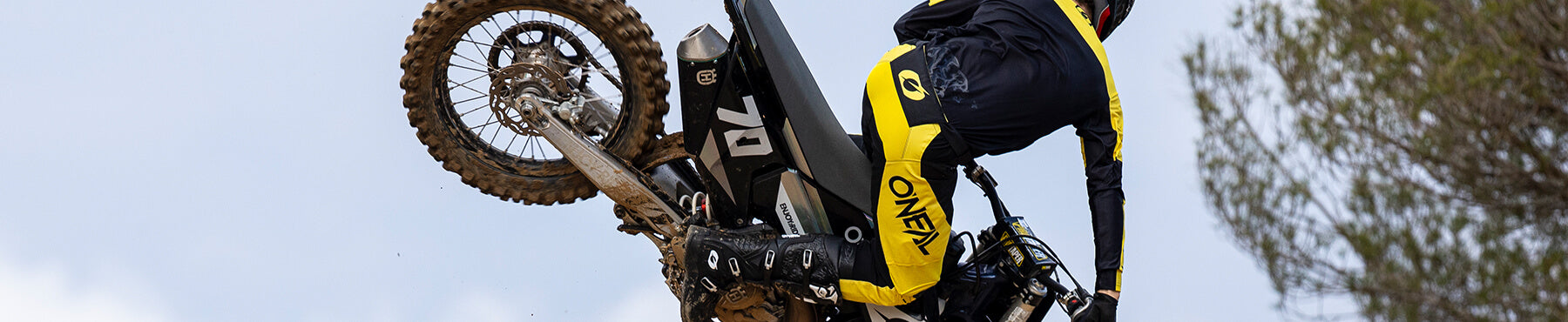 Motorradfahrer in der Luft, der einen Stunt auf einem Dirtbike vorführt. Er trägt ein gelb-schwarzes Outfit. Im Hintergrund sind ein klarer Himmel und einige Äste zu sehen.