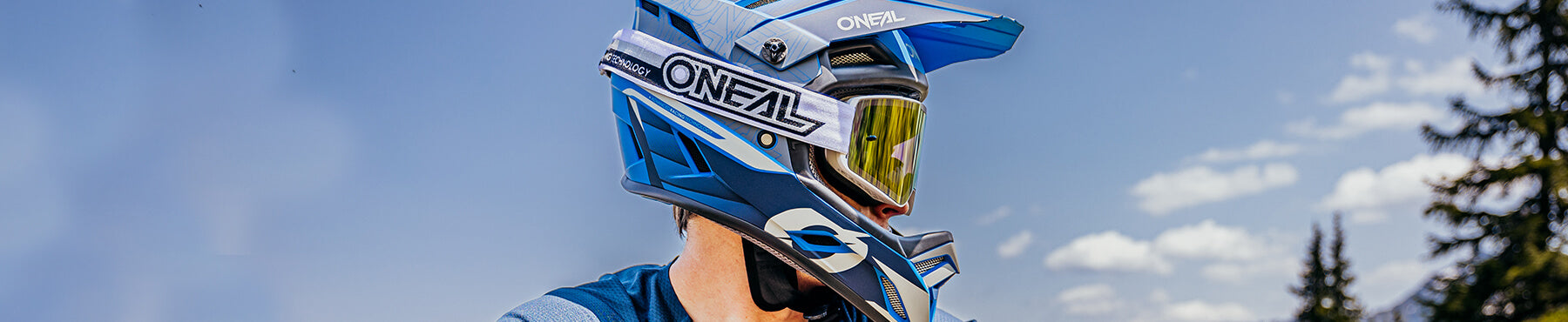 Person mit blauem O'Neal-Motocrosshelm und Schutzbrille, blickt zur Seite, vor dem Hintergrund eines blauen Himmels und von Bäumen.