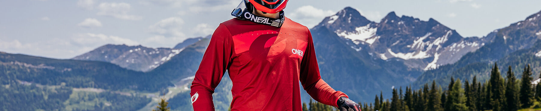 Eine Person in roter Fahrradkleidung und Helm hält ein Mountainbike auf einem Pfad. Im Hintergrund sind schneebedeckte Berge und Kiefern unter einem klaren Himmel zu sehen.