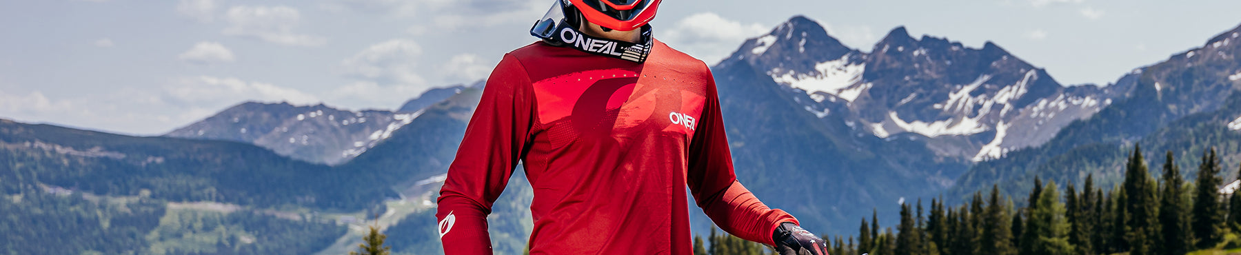 Eine Person in roter Fahrradkleidung mit Fahrradhelm und Schutzbrille steht vor einer Berglandschaft mit blauem Himmel und vereinzelten Wolken.