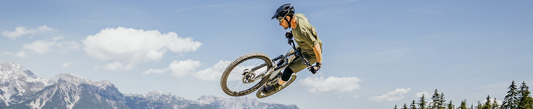 Eine Person mit Helm und Schutzausrüstung führt auf einem Mountainbike vor dem Hintergrund eines klaren blauen Himmels, Wolken und entfernter Berggipfel einen Trick in der Luft aus. Im Vordergrund sind bewaldete Hügel zu sehen.