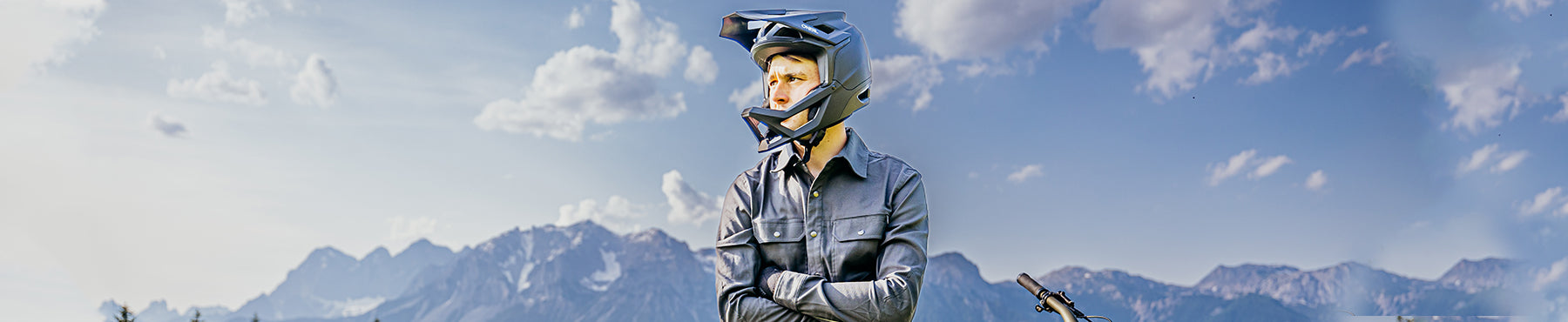 Eine Person mit dunklem Schutzhelm und Jacke steht mit verschränkten Armen da und blickt in die Ferne. Als Hintergrund dienen eine Bergkette und ein teilweise bewölkter Himmel. Ein Teil eines Fahrrads ist sichtbar.