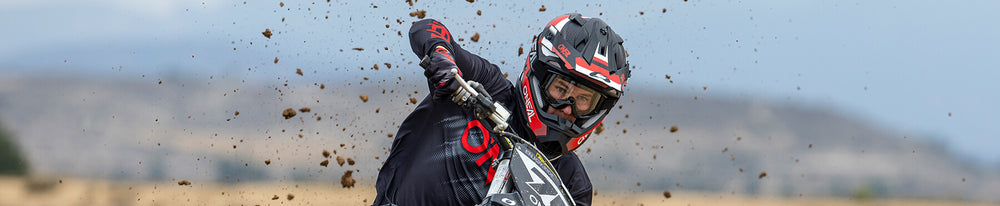 Ein Motocrossfahrer mit Helm und Schutzbrille lehnt sich in eine Kurve und wirbelt dabei Staub über die Strecke. Im Hintergrund ist eine verschwommene Landschaft mit Hügeln unter einem bewölkten Himmel zu sehen.