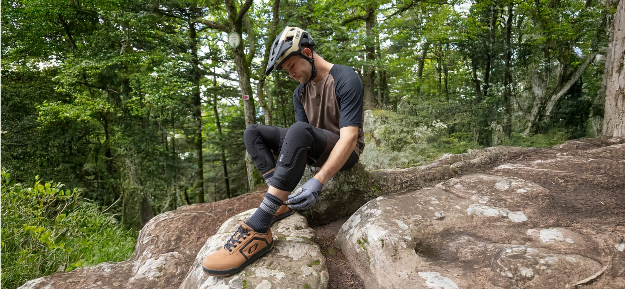 Zwei Mountainbiker fahren einen felsigen, moosbewachsenen Waldweg hinunter. Der vordere Fahrer hebt sein Vorderrad an, während der andere folgt. Das Sonnenlicht fällt durch die hohen Bäume. Der Text lautet "26 ONEAL MTB.".