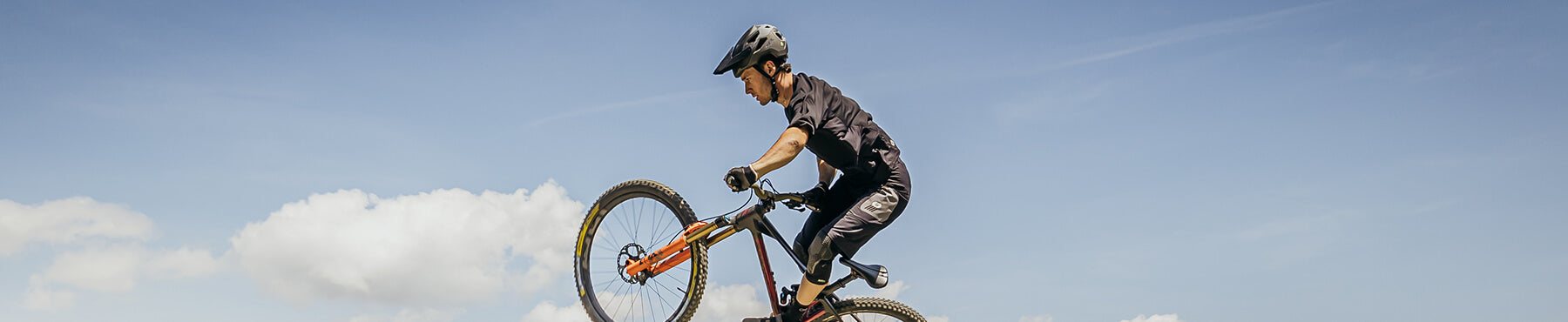 Eine Person macht auf einem Mountainbike einen Wheelie vor einem klaren blauen Himmel mit ein paar Wolken. Sie trägt einen Helm und Radsportbekleidung.