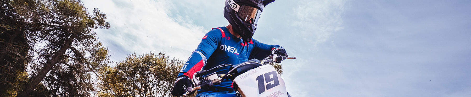 Ein Motocrossfahrer im blau-roten Outfit fährt ein Dirtbike mit der Nummer 19 auf der Vorderseite. Der Himmel ist klar und im Hintergrund sind Bäume.