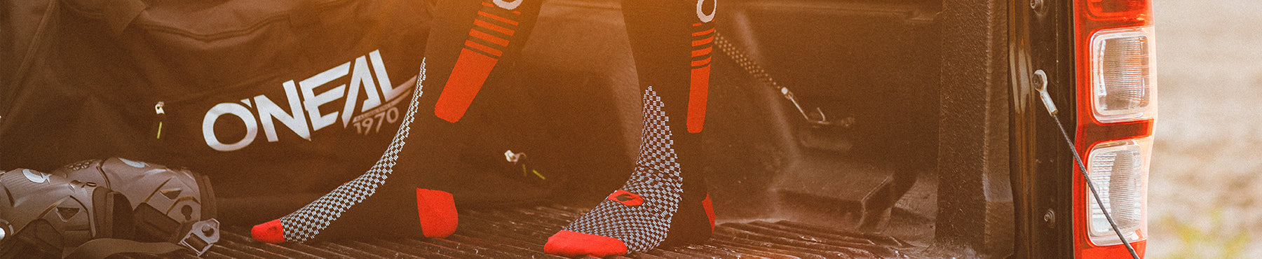 Ein Paar schwarz-rot gemusterte Socken auf der Ladeklappe eines Pickups, daneben eine schwarze Sporttasche mit der Aufschrift „ONEAL 1970“ und ein schwarzer Helm. Die Szene ist in warmes Sonnenlicht getaucht.