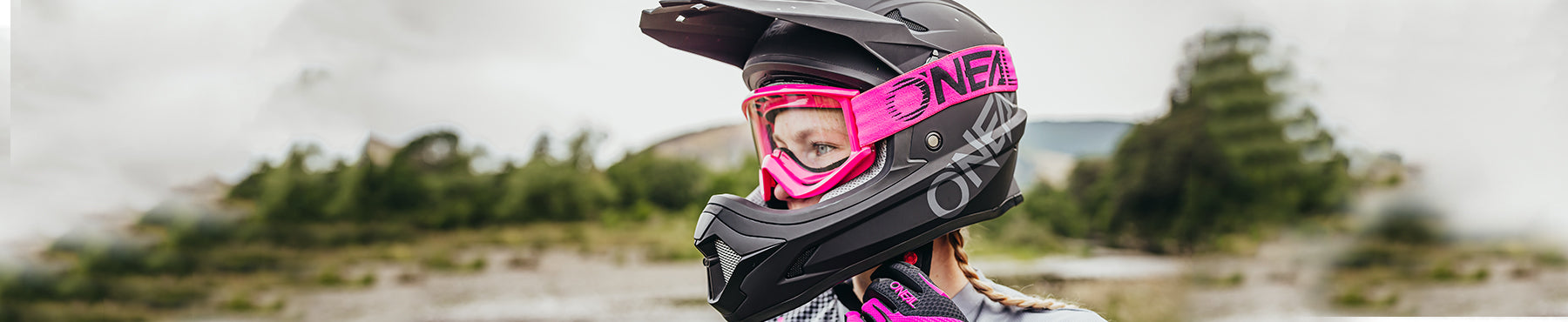 Person mit schwarzem Helm mit rosa Visier und Handschuhen, Blick nach links. Im Hintergrund ist eine natürliche Umgebung im Freien mit verschwommenen Bäumen zu sehen.