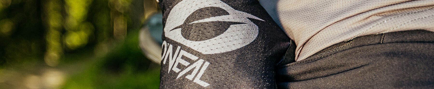 Nahaufnahme des Arms einer Person, die einen schwarzen Ärmel mit einem weißen „O'NEAL“-Logo trägt. Der Hintergrund zeigt verschwommenes Grün und Sonnenlicht.