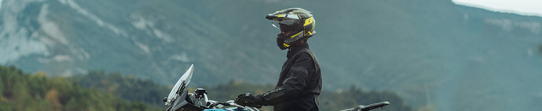 Eine Person in voller Motorradmontur und mit Helm fährt vor einer Bergkulisse Motorrad. Der Fahrer blickt nach links und das Motorrad scheint ein Tourenmodell zu sein.