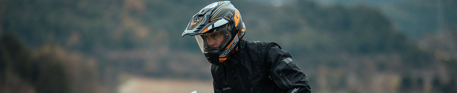 Eine Person mit einem schwarz-orangen Motorradhelm und einer schwarzen Jacke steht im Freien mit verschwommener Natur im Hintergrund.