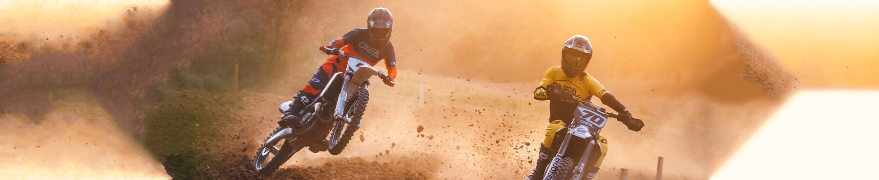 Zwei Motocrossfahrer fahren auf einer Sandpiste ein Rennen. Einer trägt Rot und Schwarz, der andere Gelb. Um sie herum wirbelt Staub auf, und das Sonnenlicht erzeugt einen warmen Schein im Hintergrund.