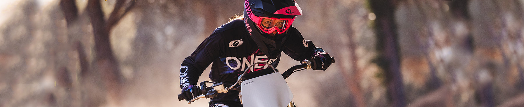 Eine Person mit einem rosa Helm und schwarzer Motocross-Ausrüstung fährt mit einem Dirtbike durch ein Waldgebiet. Hinter ihr wirbelt Staub auf, während sie sich auf den vor ihr liegenden Weg konzentriert.