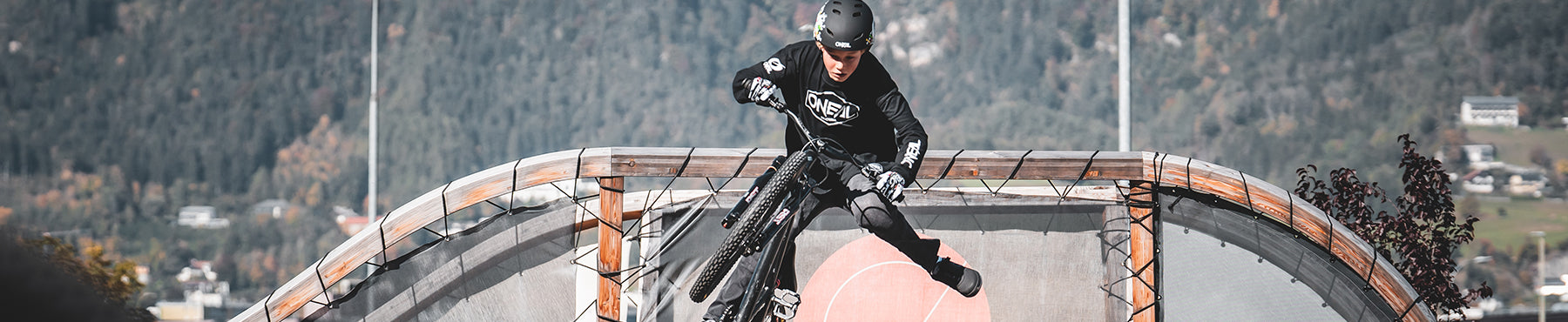 Ein Radfahrer mit Helm und Schutzausrüstung führt auf einer Rampe in einem Skatepark einen Trick vor. Im Hintergrund sind eine Berglandschaft und einige Gebäude zu sehen.