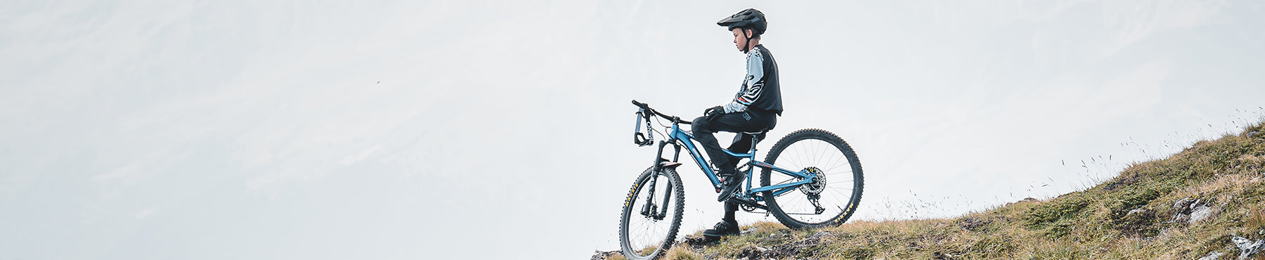 Eine Person mit Helm sitzt auf einem Mountainbike am Rand eines grasbewachsenen Hügels und blickt auf eine neblige Landschaft.