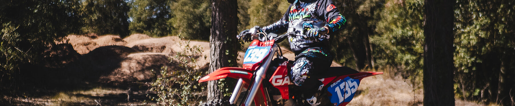 Ein Motocrossfahrer in bunter Montur rast auf einem roten Dirtbike mit der Nummer 158 durch ein Waldstück. Im Hintergrund sind Bäume und Erdhügel unter einem klaren Himmel zu sehen.
