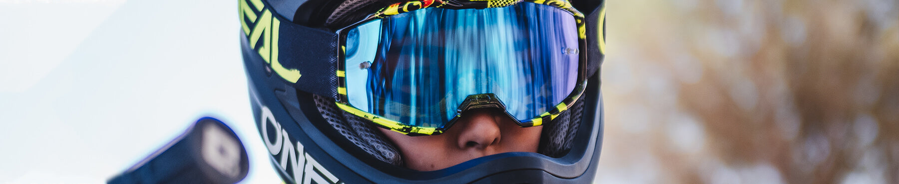 Eine Person trägt einen Helm und eine reflektierende Schutzbrille, die eine verschwommene Außenumgebung zeigt. Auf dem Helm ist eine fettgedruckte Beschriftung angebracht und das Gesicht der Person ist teilweise durch die Schutzbrille sichtbar.