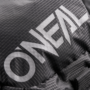 Close-up of the O'NEAL JUNCTION Protective Backpacks from O'NEAL Europe. You can see a black, structured fabric with thin white grid lines and the print "O'NEAL" in big, bold light gray letters. The material is slightly wrinkled.