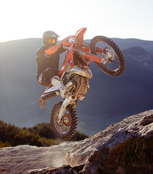 Ein Motocrossfahrer in schwarzer Ausrüstung und mit Helm vollführt auf einem robusten roten Dirtbike auf felsigem Gelände einen waghalsigen Wheelie. Im Hintergrund ist eine Berglandschaft zu sehen, und die untergehende Sonne wirft einen warmen Schein auf die Szene.
