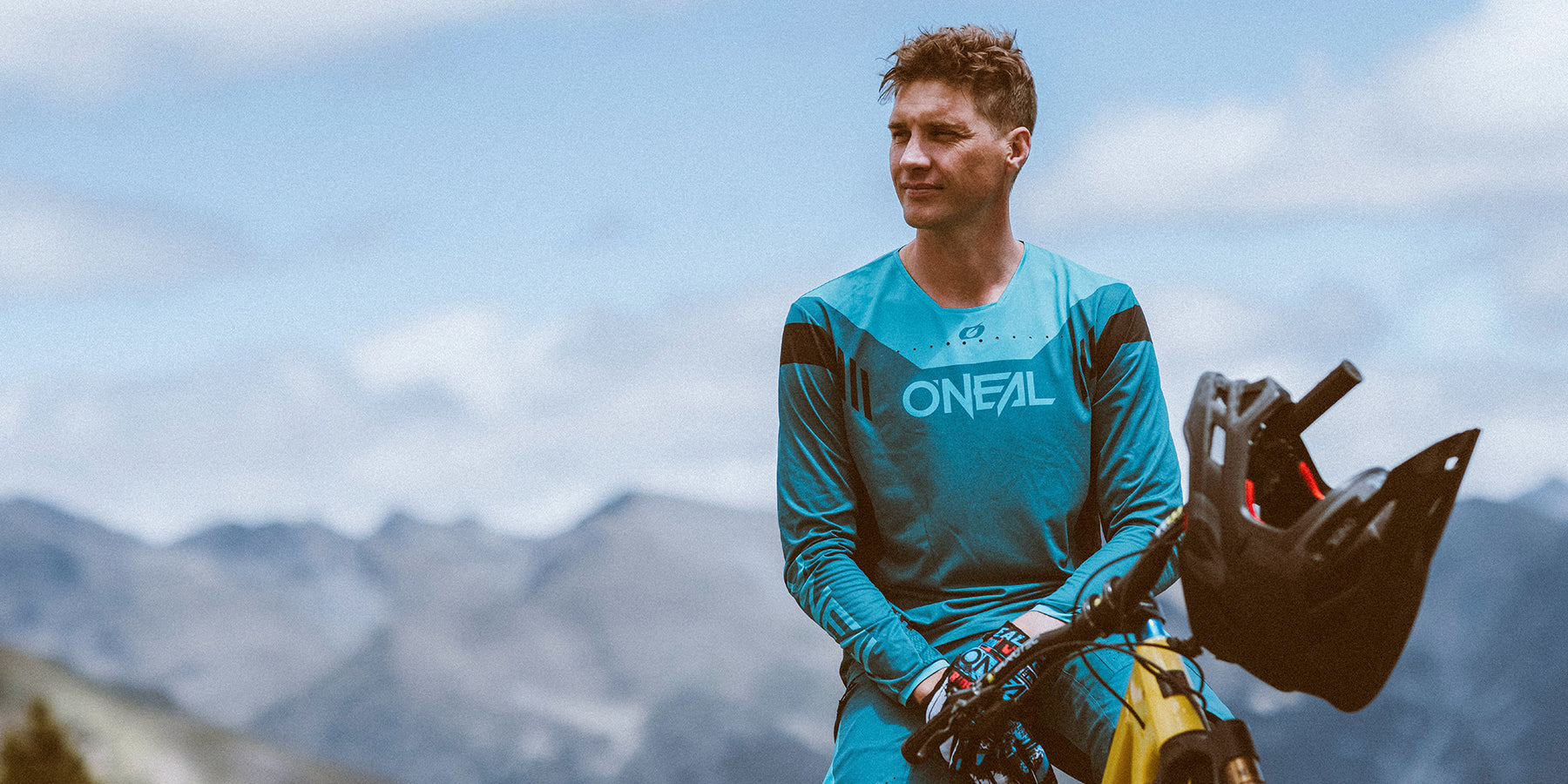 Eine Person im blauen ONFAL-Radsport-Outfit steht im Freien neben einem Mountainbike. Sie hält einen schwarzen Helm unter dem Arm und blickt in die Ferne. Im Hintergrund sind Berge und ein teilweise bewölkter Himmel zu sehen.