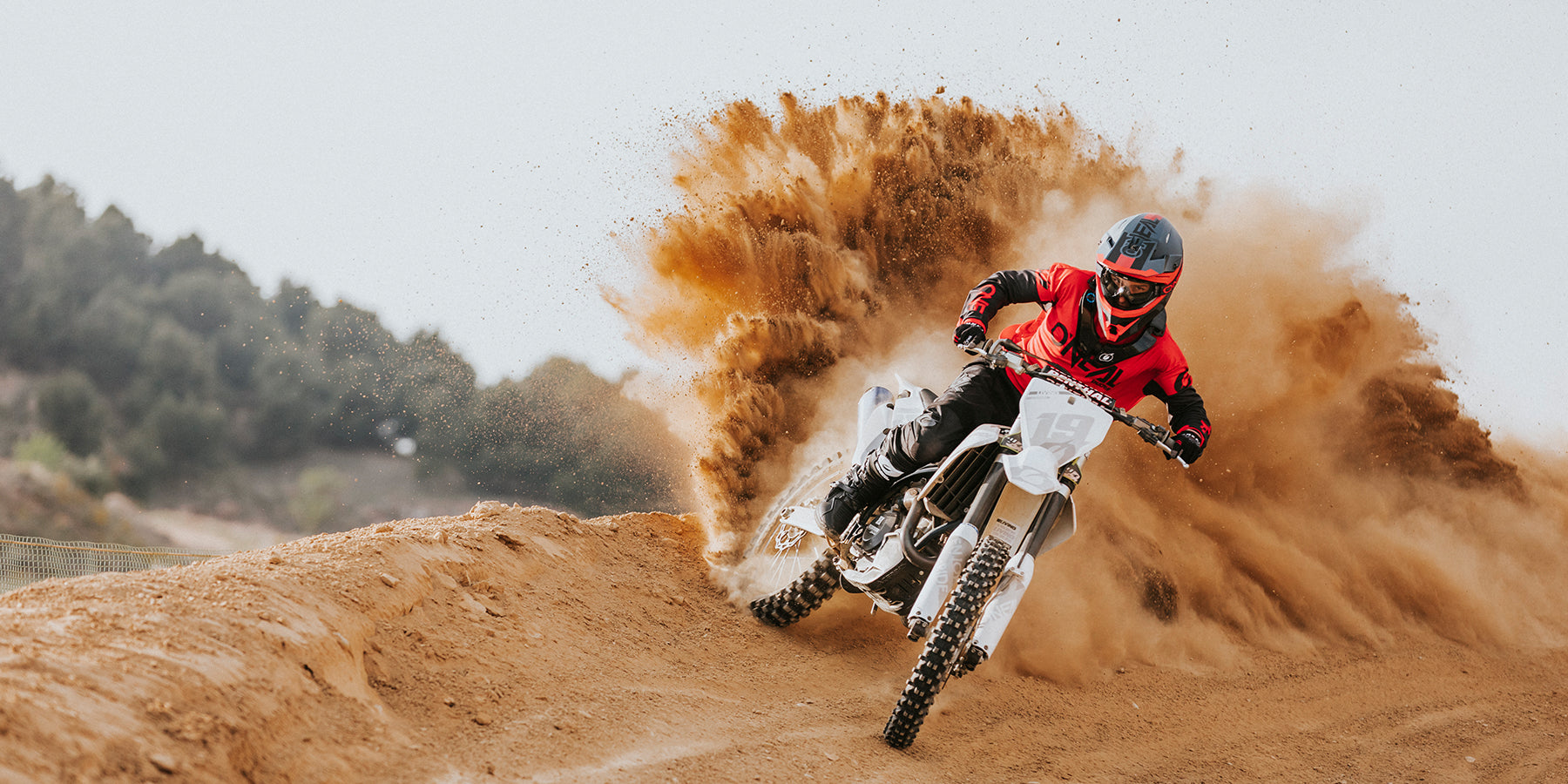 Ein Motocrossfahrer in rot-schwarzem Outfit und Helm fährt aggressiv eine Kurve auf einer unbefestigten Strecke, wodurch eine große Staubwolke hinter ihm in die Luft geschleudert wird. Im Hintergrund sind Bäume und ein dunstiger Himmel zu sehen.