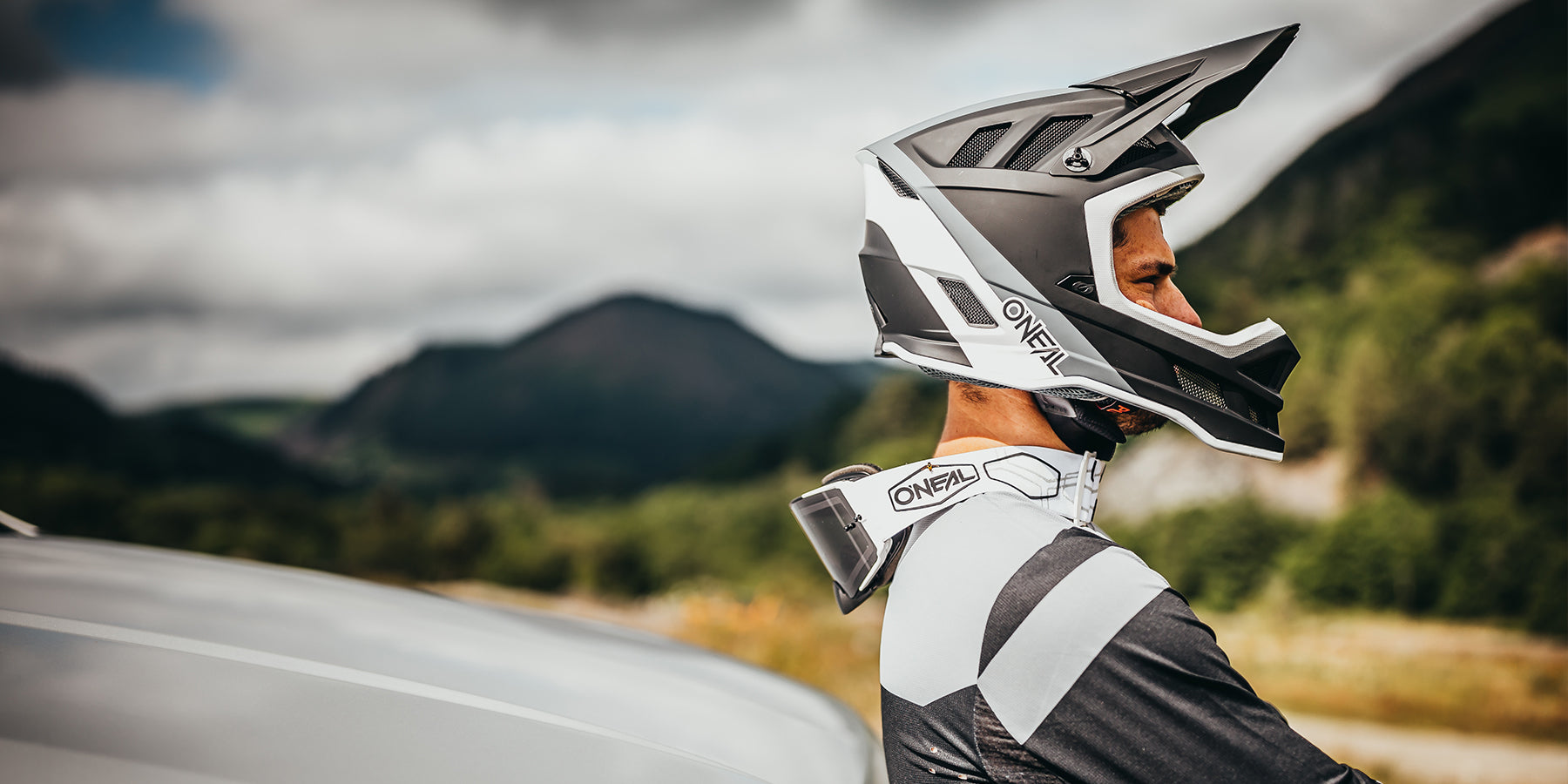 Eine Person mit einem schwarz-weißen Integralhelm und passender Motocross-Ausrüstung steht neben einem Fahrzeug und blickt in die Ferne mit einer Berglandschaft und einem bewölkten Himmel im Hintergrund. Die Ausrüstung ist mit „ONEAL“-Logos versehen.
