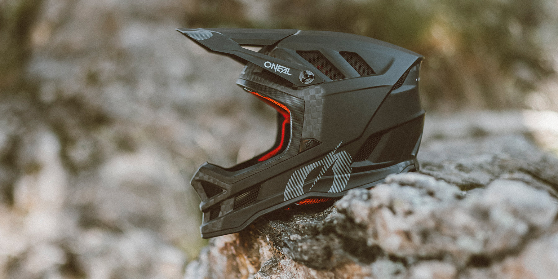 Ein schwarz-grauer O'Neal-Integralhelm mit Visier und roter Innenpolsterung thront auf einem Felsen vor einem verschwommenen natürlichen Hintergrund. Der Helm zeichnet sich durch ein stromlinienförmiges Design mit Belüftungsöffnungen und auffälligem Branding aus.