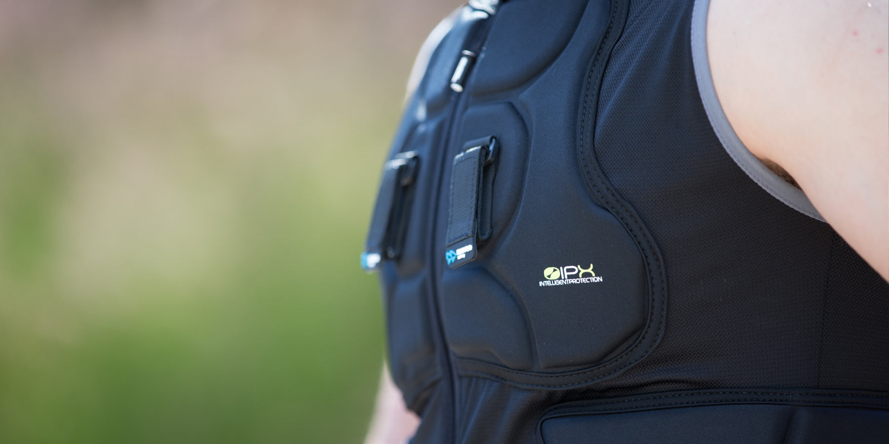 Nahaufnahme einer Person, die eine schwarze Neoprenweste mit sichtbaren Nähten und Aufnähern mit der Aufschrift „IPX“ trägt, die zum Aufprallschutz dient. Der Hintergrund ist verschwommenes Grün. Die Person trägt unter der Weste ein ärmelloses Hemd.