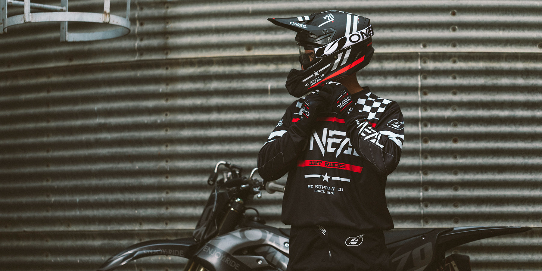 Eine Person mit Integralhelm und Motocross-Ausrüstung steht neben einem geparkten Dirtbike. Der Hintergrund besteht aus gewelltem Metall, was ein industrielles Gefühl vermittelt. Die Person scheint den Helmgurt zu regulieren und sich auf eine Fahrt vorzubereiten.