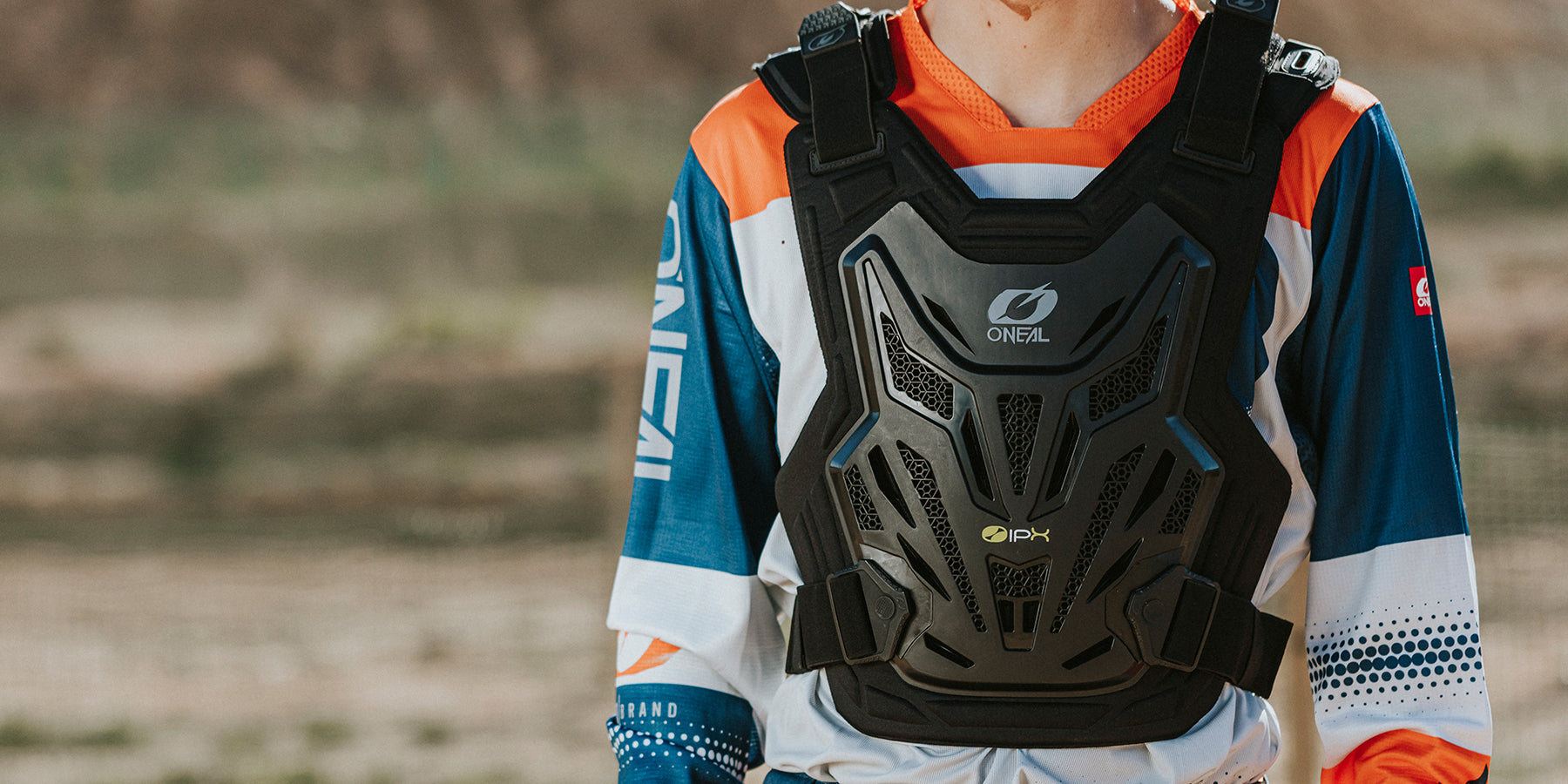 Eine Person, die eine Brustschutzweste und ein buntes Motocross-Trikot trägt, steht im Freien. Die Weste mit der Marke „ONEAL“ schützt den Oberkörper, während das blau-orange-weiße Trikot mit Texten und Logos versehen ist. Der Hintergrund ist verschwommenes Gelände.