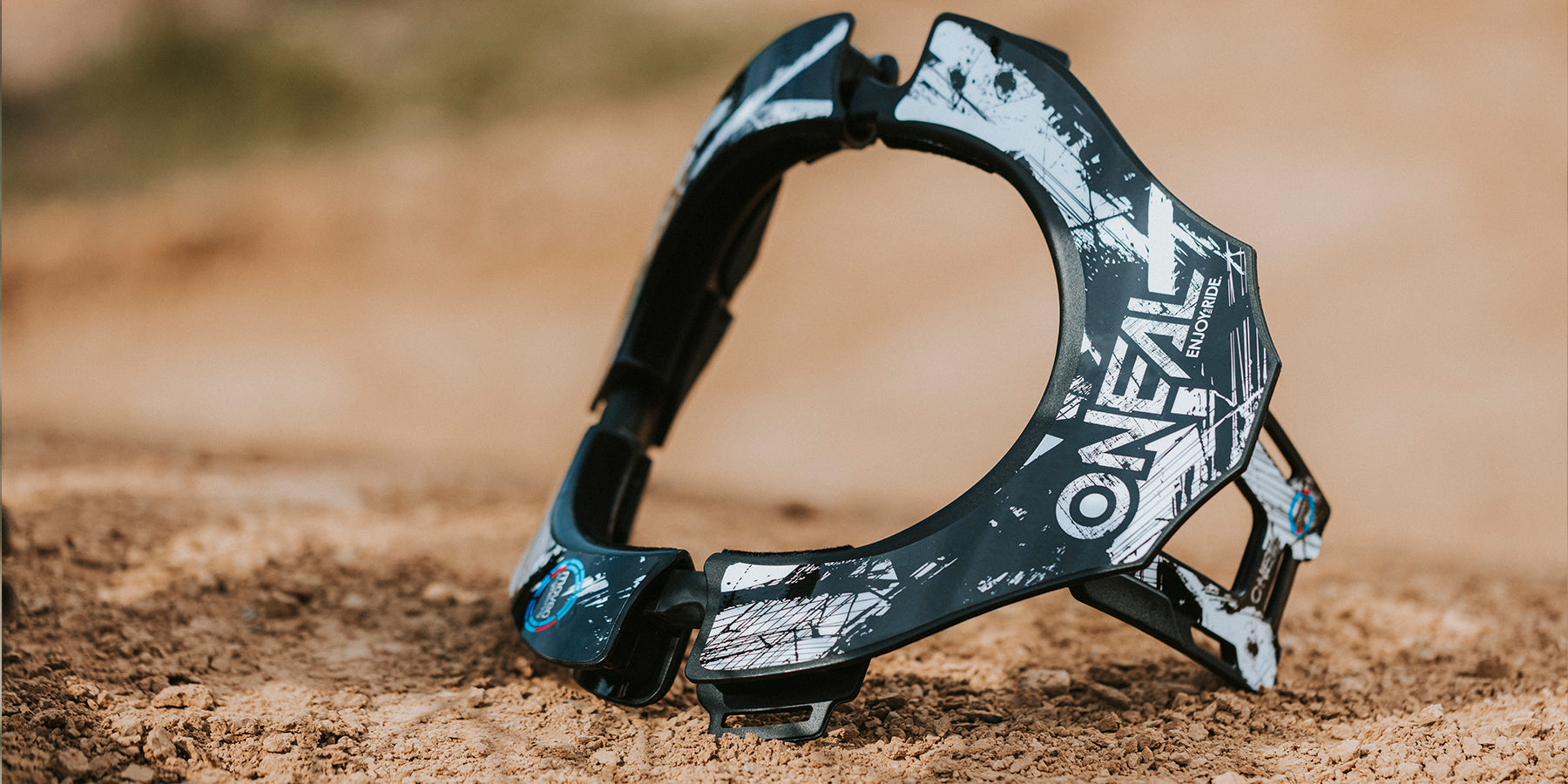 Eine schwarz-weiße O'Neal-Halskrause liegt auf sandigem Untergrund. Die Schutzausrüstung, die für Offroad- und Motocross-Fahrer entwickelt wurde, weist ein robustes, grafisches Design auf, auf dem der Markenname deutlich zu sehen ist. Der Hintergrund ist verschwommen, wodurch die Details der Krause hervorgehoben werden.