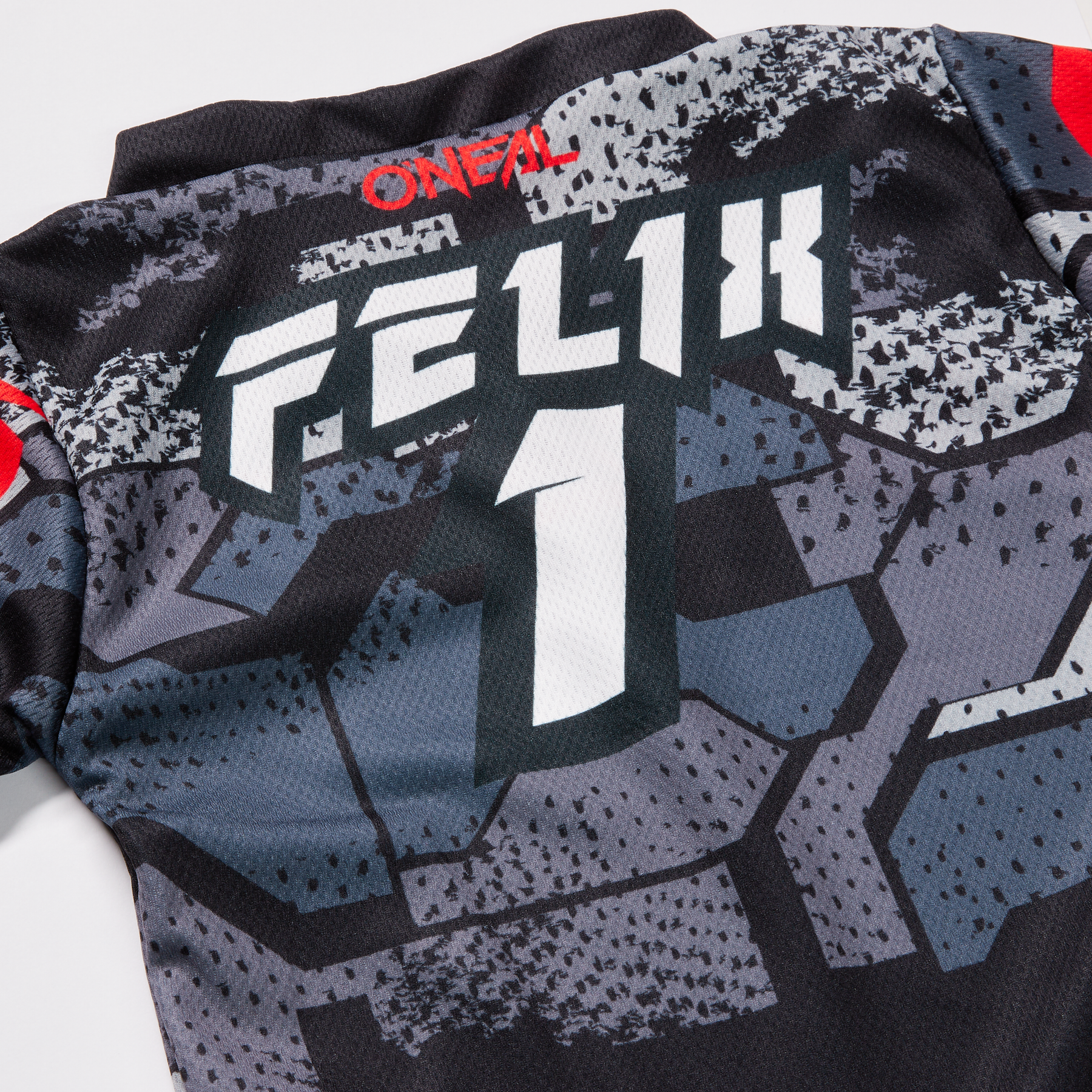 Nahaufnahme der Rückseite eines schwarz-grauen O'NEAL Custom Jersey Kinder CAMO Sporttrikots mit dem Text „Felix 1“ in fettem, weißem Druck. Das im Sublimationsverfahren hergestellte Shirt trägt oben außerdem den Markennamen O'NEAL in Rot. Das Design besticht durch große, geometrische Muster in Schwarz und Grautönen für einen einzigartigen Look.