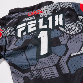 Nahaufnahme der Rückseite eines schwarz-grauen O'NEAL Custom Jersey Kinder CAMO Sporttrikots mit dem Text „Felix 1“ in fettem, weißem Druck. Das im Sublimationsverfahren hergestellte Shirt trägt oben außerdem den Markennamen O'NEAL in Rot. Das Design besticht durch große, geometrische Muster in Schwarz und Grautönen für einen einzigartigen Look.