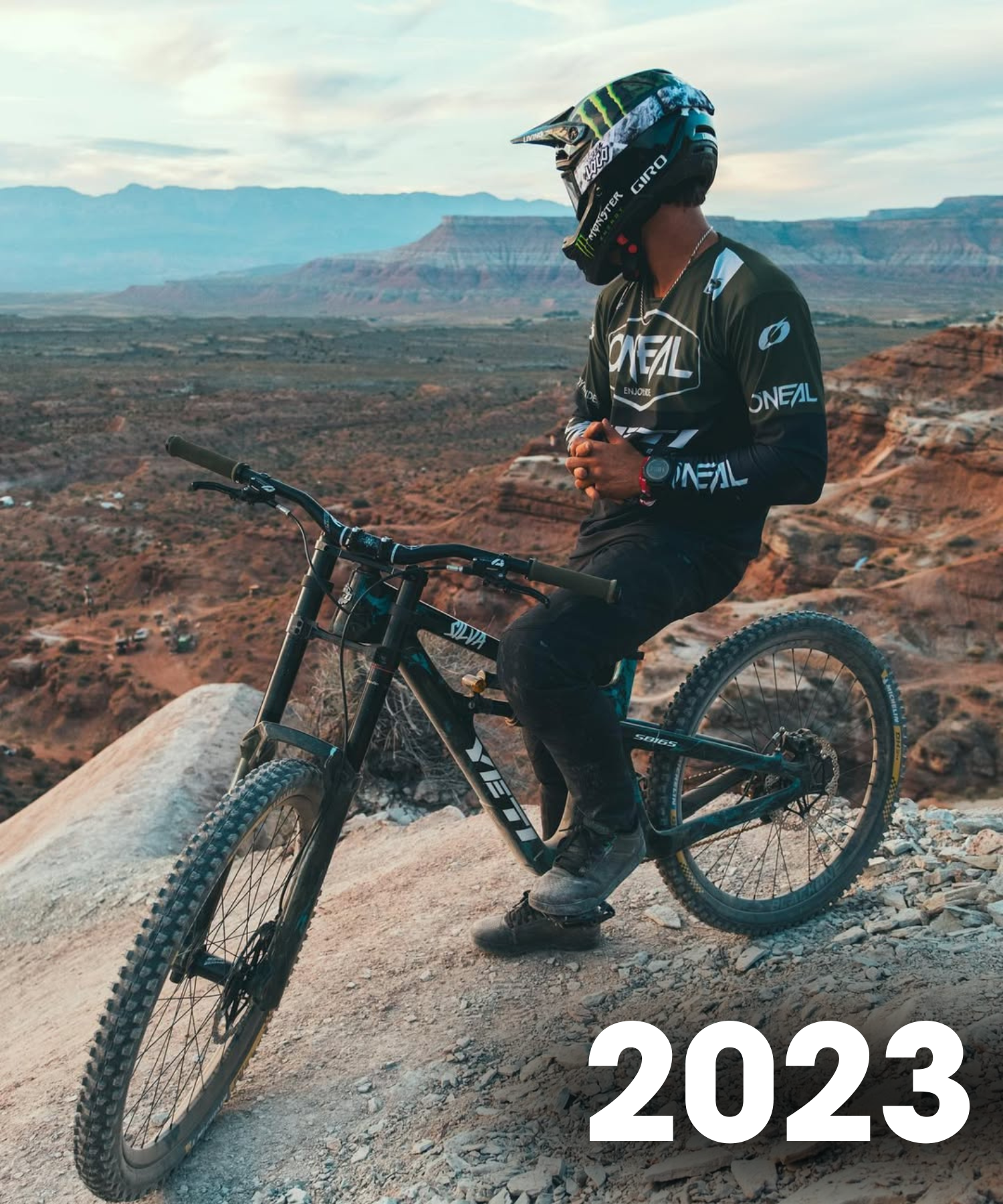 Ein Mountainbiker in voller Montur und mit Helm sitzt auf einem schwarzen Fahrrad am Rande einer felsigen Klippe und blickt auf eine weite Wüstenlandschaft. Die Zahl "2023" ist in weißer Schrift in der unteren rechten Ecke zu sehen.