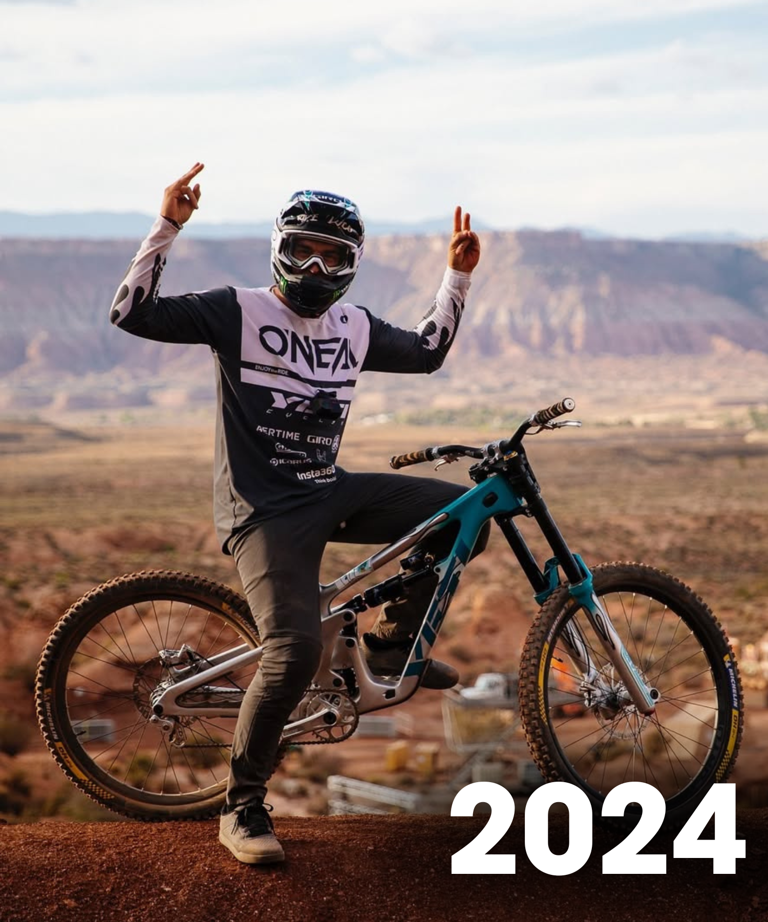 Ein Mountainbiker in voller Montur posiert mit Peace-Zeichen, während er auf einem Fahrrad an einem Aussichtspunkt sitzt, mit "2024" in fetter weißer Schrift unten rechts.
