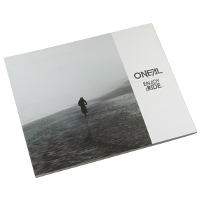 Das geschlossene O'NEAL ENJOY THE RIDE BOOK zeigt eine einsame Figur an einem nebligen Strand, die Emotionen hervorruft. Auf der rechten Seite ist das O'NEAL-Logo und der Schriftzug "ENJOY THE RIDE." in Schwarz auf Weiß zu sehen. Marke: O'NEAL. Produkt: O'NEAL ENJOY THE RIDE BOOK (KOSTENLOSER GOODIE-WAGEN).